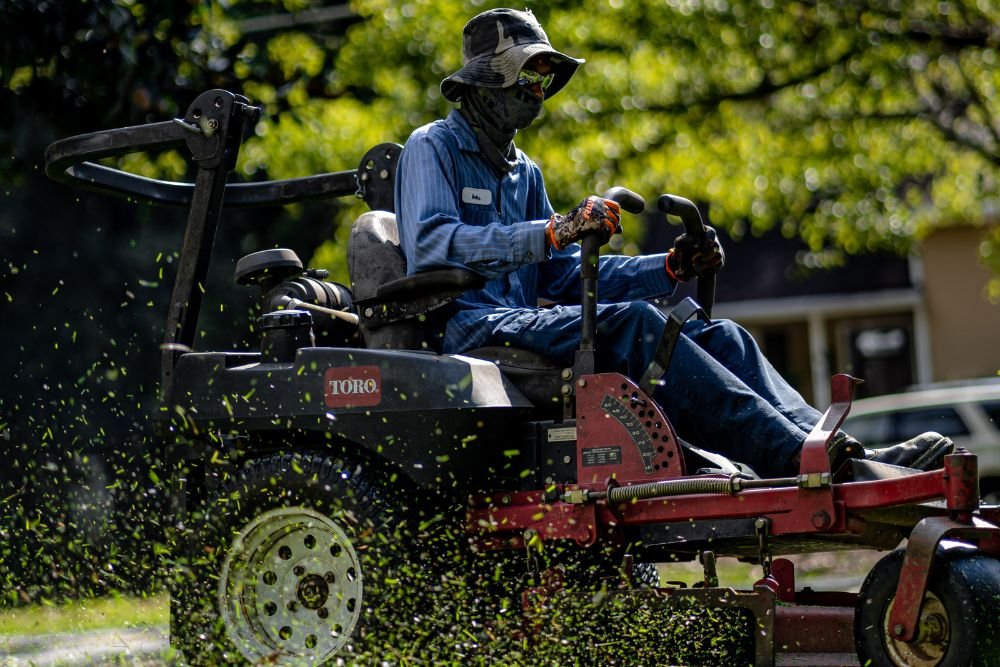 A man uses a lawn mower and holds a leaf blower, tending to a neat lawn surrounded by trees.