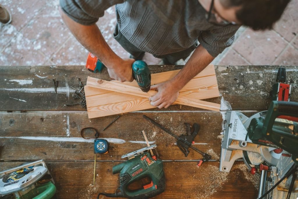 A man diligently works at a wooden table, surrounded by various tools, focused on his task.