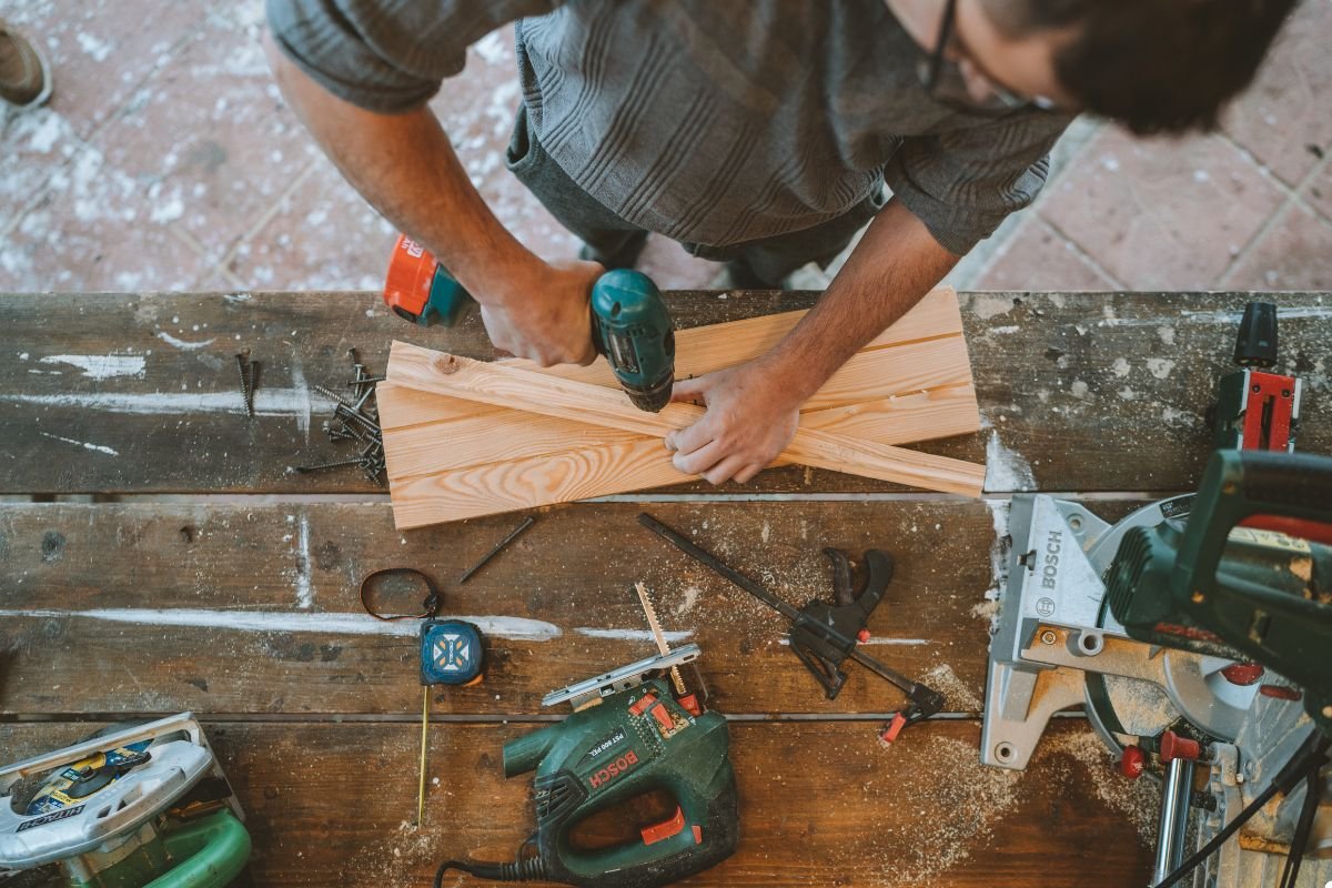 A man diligently works at a wooden table, surrounded by various tools, focused on his task.
